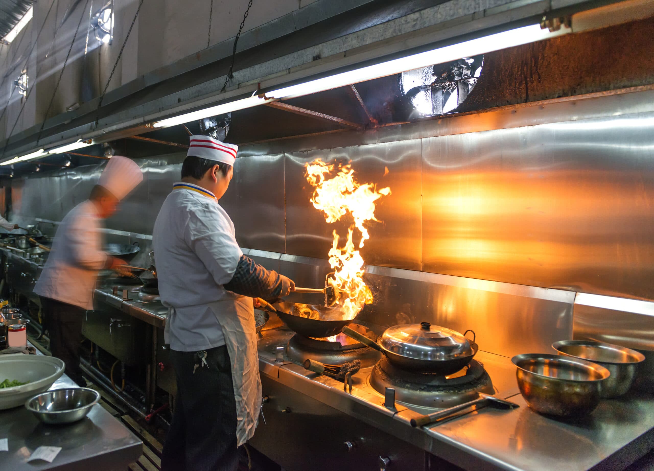 Chefs cooking over open flame beneath a commercial kitchen exhaust hood