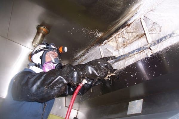 Technician in a respirator scraping grease from a commercial kitchen hood plenum